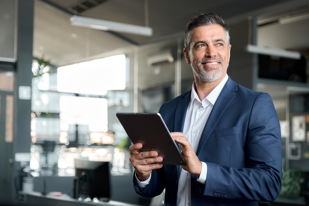 Happy middle aged business man ceo wearing suit standing in office using digital tablet. Investment property builders